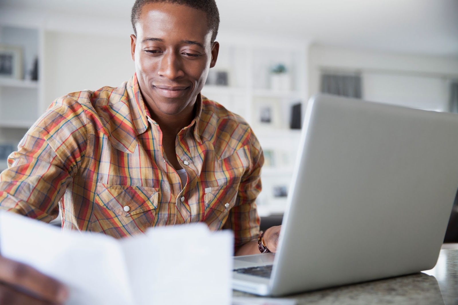 Imagem de um homem negro, de cabelos curtos e vestindo uma camisa xadrez. Ele sorri enquanto analisa um papel em sua mão direita, enquanto a esquerda está apoiada em um notebook cinza.