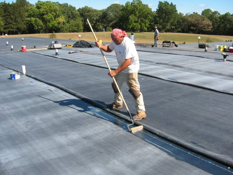 A man in a white t-shirt and a red bandana on a flat rooftop using a large paint roller to apply a waterproof coating.
