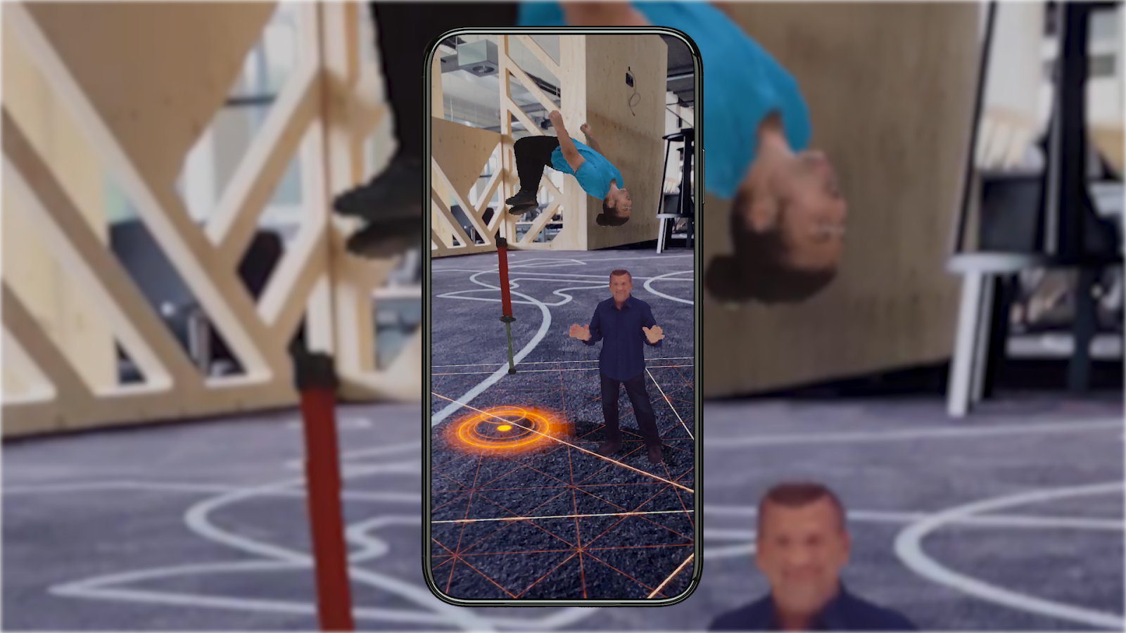 In the foreground, a mobile phone displays the scene of a man backflipping through the air, with another man standing on the ground to his right. In the background, this same image is seen, but blurred out.