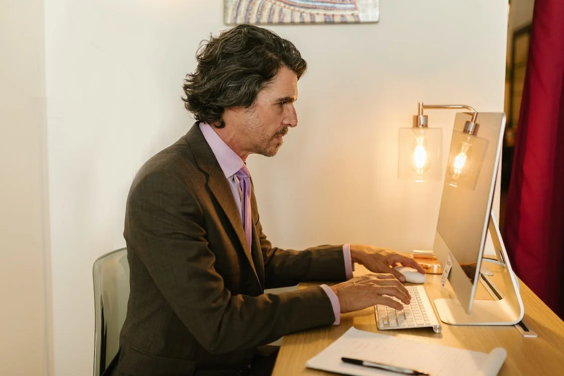 A Man in Gray Suit Typing while Looking at the Monitor
