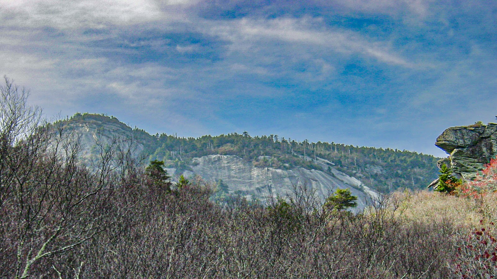 Exposed rocks against the cloudy blue sky with barren foliage in the foreground. A rock formation is on the right mid-ground. 