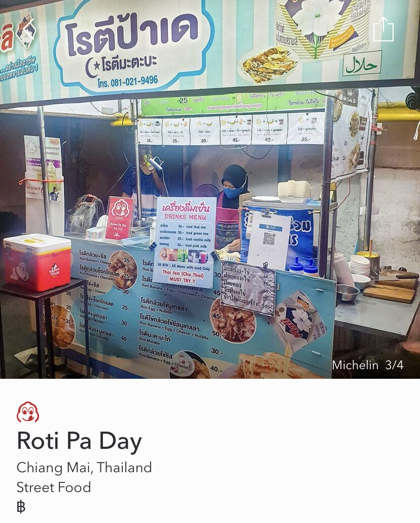 Screenshot from Michelin app, of Roti Pa Day street stall in Chiang Mai, with vendor working behind. Several menu signs on top, bottom and sides of stall.