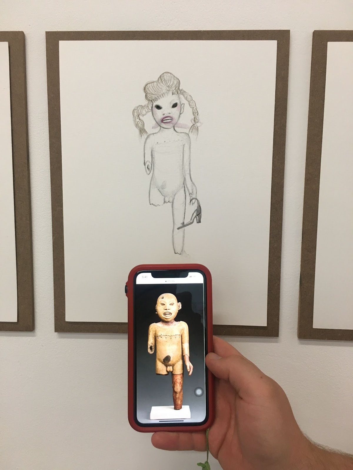 Image: A close up shot of a figure drawing by Sofia Moreno. The figure has a broken leg and is carrying a heeled shoe. In front of the drawing extends a hand holding a phone, which shows an image from the internet of an ancient figurative sculpture to draw comparison. Photo by Noa/h Fields.