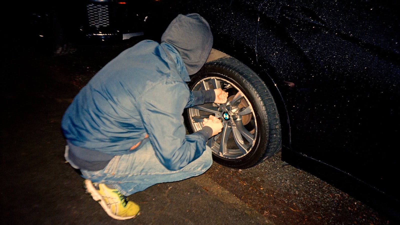 A hooded activist kneels beside a parked SUV 
