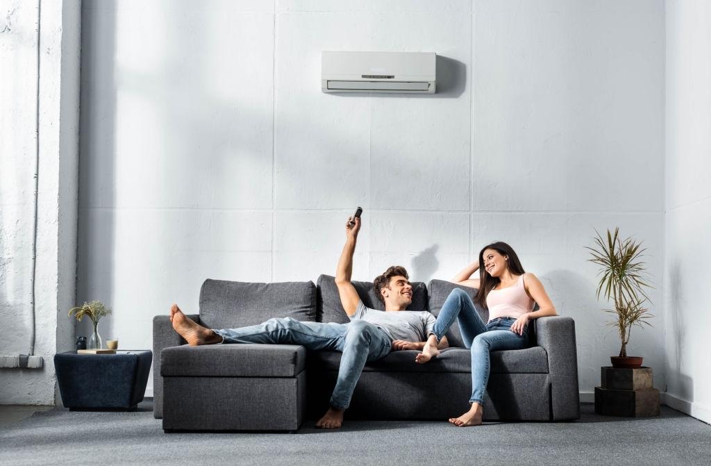 WeServe | A woman and man sitting on a dark grey couch smiling with the man raising his arm turning on the AC with a white background wall and grey carpet 