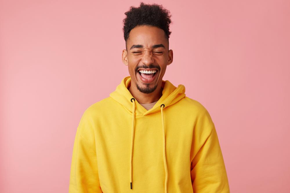 Young happy smiling african american guy in yellow hoodie, heard a very funny joke and laughed, standing on a pink background with eyes closed.