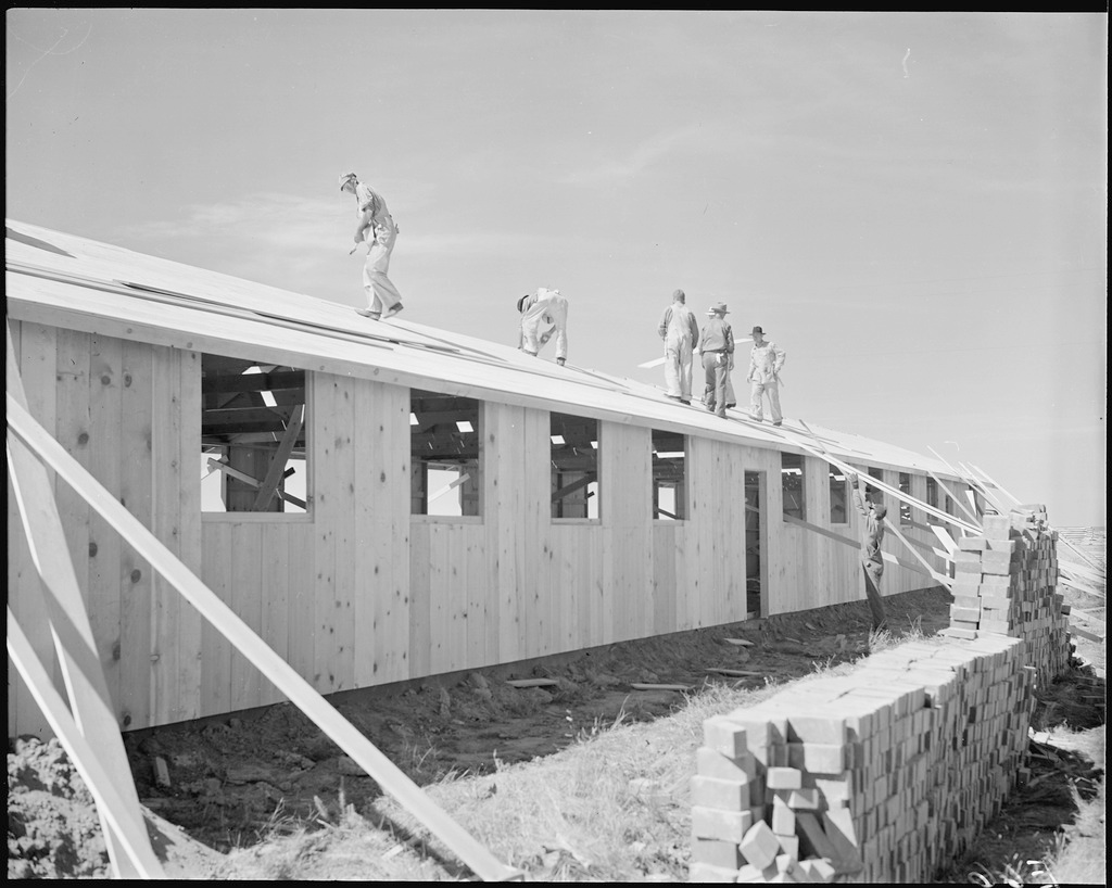 Construction workers roofing an assembled barracks unit in Amache concentration camp. Five men are working on the roof and another is passing up a plank of wood. Bricks are stacked on the ground next to the unfinished building.