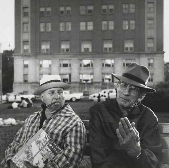 A black and white image of 2 men sat on a bench having a discussion by Vivian Maier