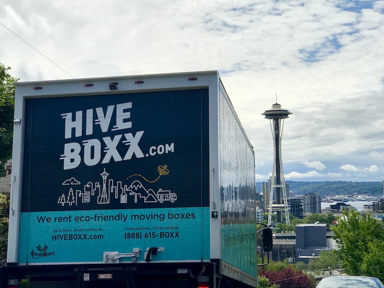 A truck going down a street with advertisement on the back of it saying 'hive boxx.com' and ' We rent eco-friendly moving boxes' 