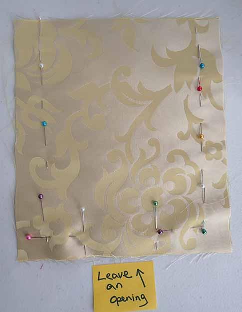 Two pieces of a cream silk fabric with a floral design pinned together with an opening on the bottom edge. A sticky note has an arrow pointing at the opening an the note "leave an opening" below the fabric.