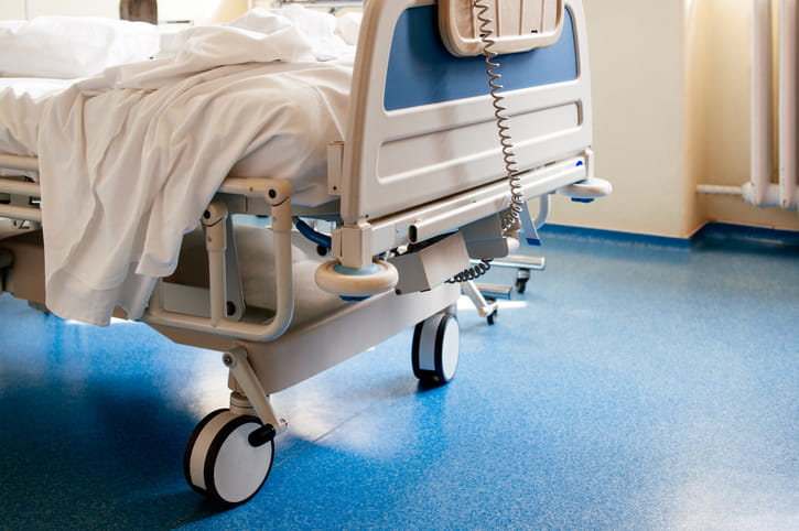 Lower half of a white and blue hospital bed used for medical care with the blankets ruffled. The lower half of the photo shows the hospital wheels against a blue hospital floor.