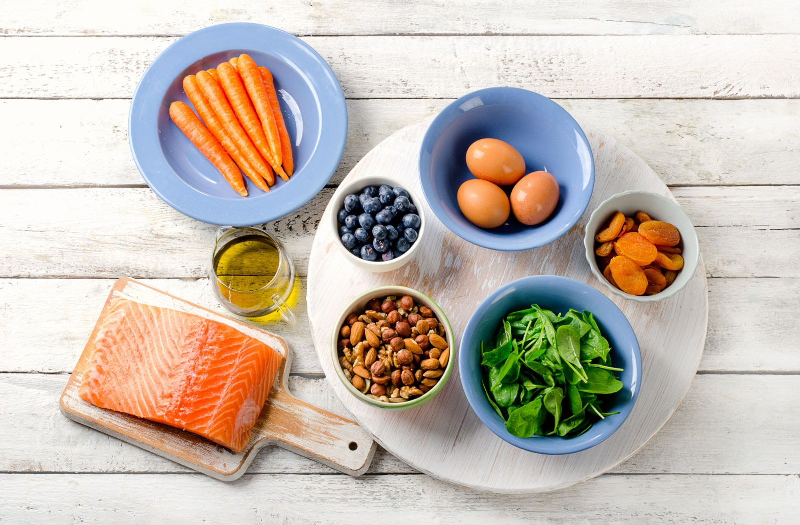 A variety of eye-healthy foods sitting out on a table, including salmon, blueberries, nuts, spinach, dried apricots, eggs, carrots and oils.