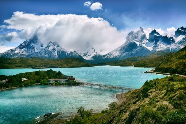 A bridge over turquoise blue water leading to charming building nestled in lush greenery with the backdrop of snow capped mountains found in Torres del Paine national park in Chile