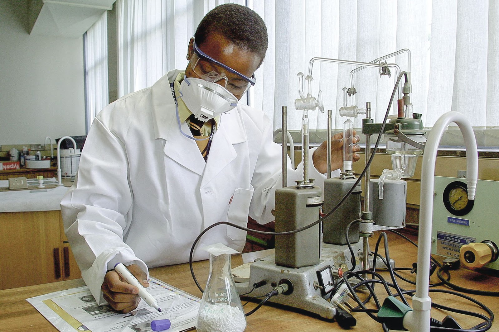Black man in lab coat highlights paper beside a test machine. 