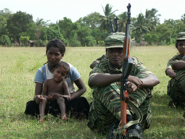 A small child and a woman sit next to LTTE cadres training in a public playground in Kilinochchi, a district in the Northern Province, in this picture taken in June 2004. The Tigers held sway over all aspects of life in areas they controlled until their defeat in 2009. Credit: Amantha Perera/IPS