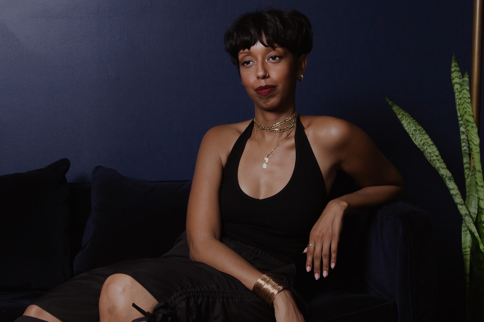 Image: The portrait features Barédu sitting upright on a dark blue couch in front of a dark blue wall. Behind her, there is a gold lamp stand and a snake plant She has on a black top and black pants and is wearing gold necklaces, bracelets, and earrings. Image captured and edited by Joshua Johnson.