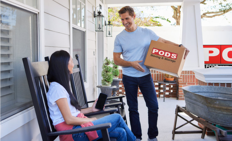 A mature couple smiles at each other on their front porch. The woman is sitting on a rocking chair, holding her phone and the man is in the process of taking a moving box to the PODS container in their driveway.