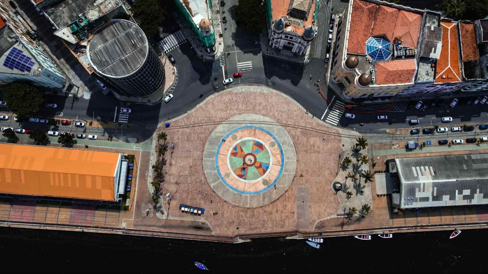 Vista aérea do Marco Zero, na Praça Rio Branco, Recife Antigo. No centro da praça, está um mosaico circular colorido. Ao redor dela, aparece uma avenida que contorna o espaço e várias construções antigas.