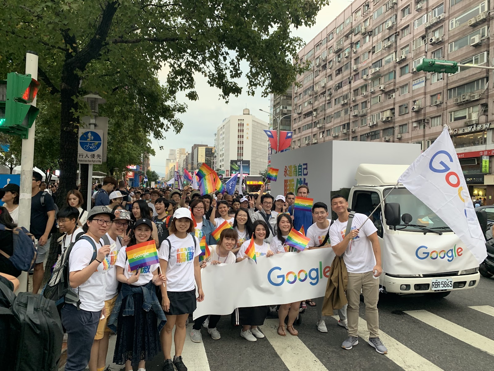 Image 1:一群參加台北同志遊行的 Google 員工，身穿印有 Google 標誌的白色 T 恤，手持彩虹旗和 Google 旗幟，背景是高樓大廈和一輛印有 Google 標誌的卡車