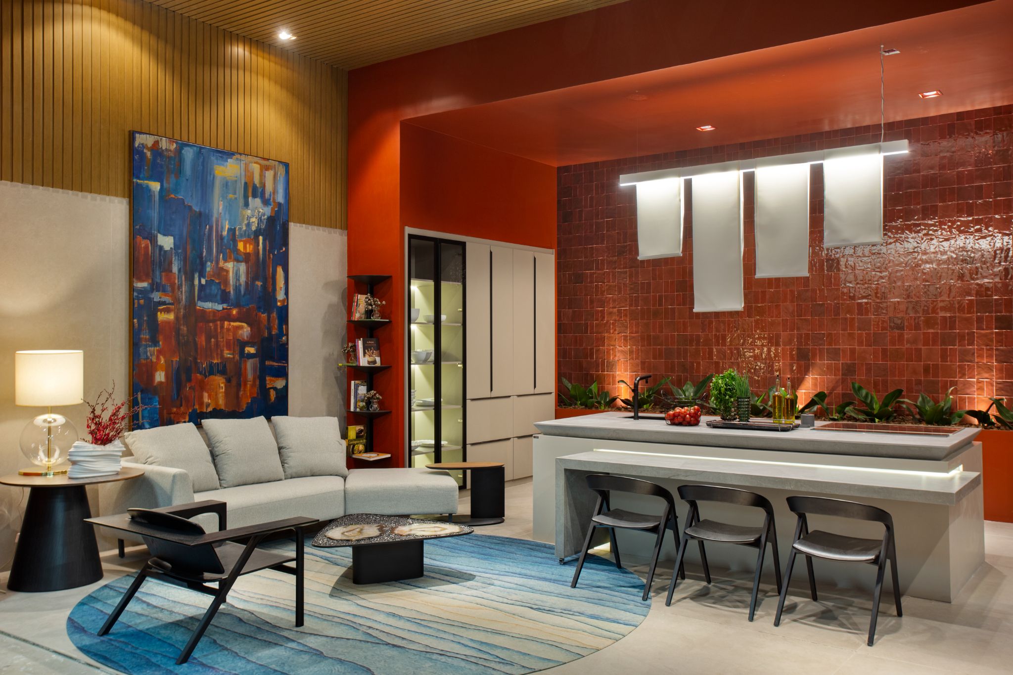 Sala de estar integrada a cozinha com parede revestida de cerâmica vermelha, bancada cinza, sofá cinza, cadeiras pretas e tapete redondo azul.