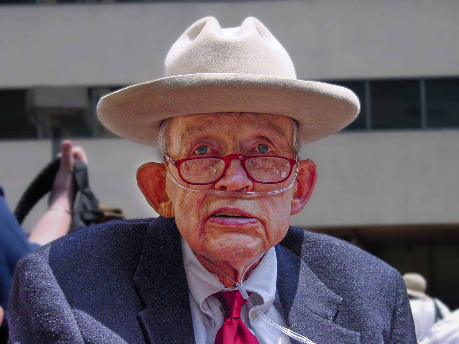 Man with sorrowful blue eyes looks over his red glasses is wearing a cowboy hat, blue suit and red tie. Oxygen tubes run into his nose.