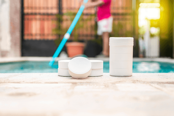 Pool chemicals sitting beside a pool, a person is cleaning the pool in the background