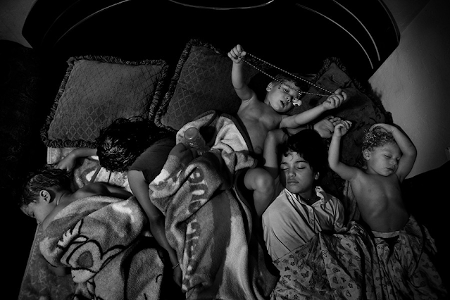 Cinco crianças dormem amontoadas em uma cama de casal. Aparentemente desconfortáveis, em razão do pouco espaço disponível. Duas das crianças dormem curvadas para a esquerda e as outras dormem de barriga para cima com os braços levantados. Na cama também há três almofadas escuras.