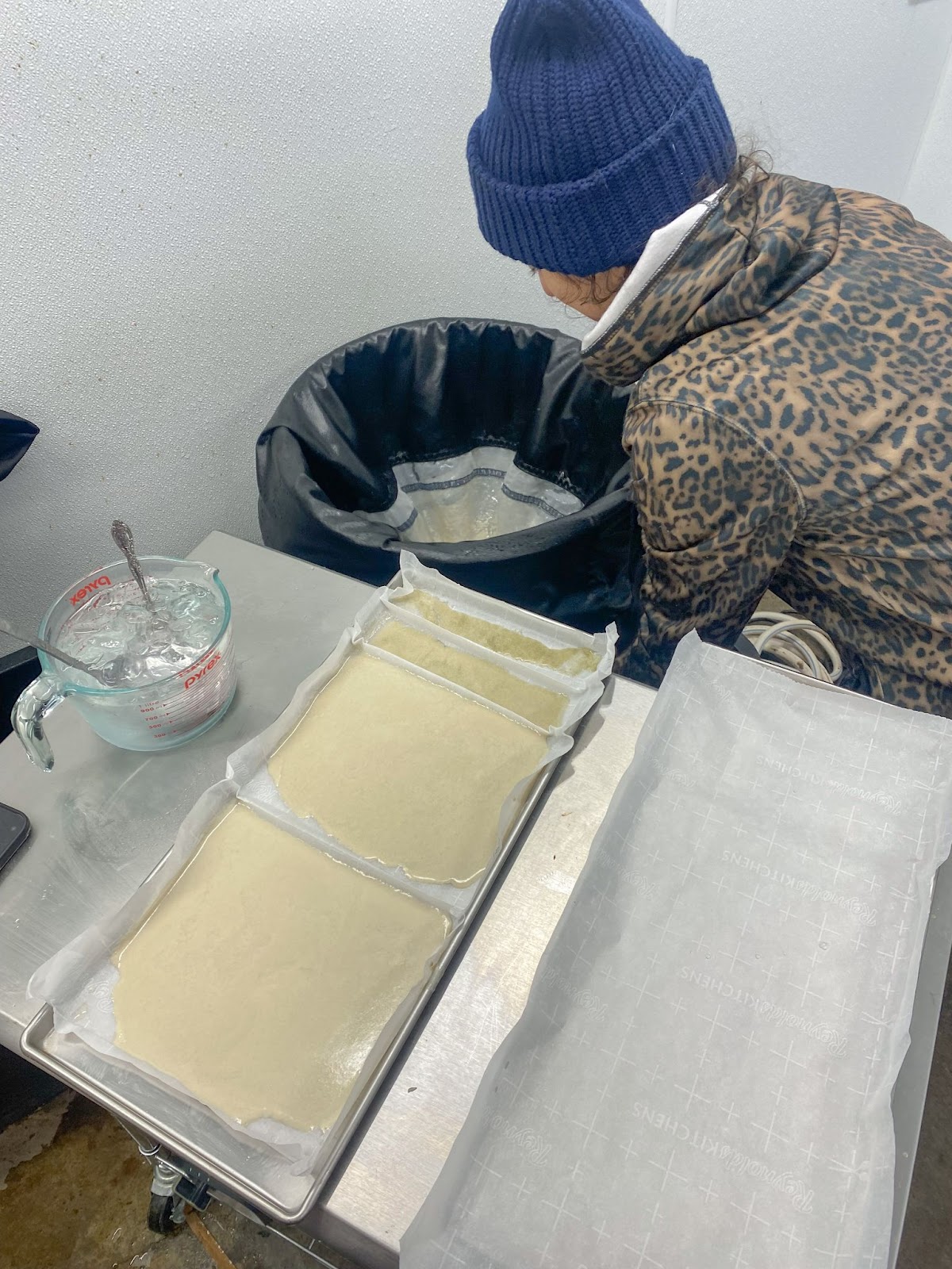 Foto colorida da Alice  coletando coletando e preparando o haxixe para secagem, com bandejas de inox, papel manteiga  e haxixe molhado para secagem