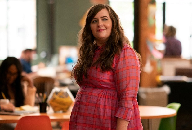 Annie in Shrill. Image: Annie stands center-frame in a pink and purple plaid dress that buttons. I tis high collared, and long sleeved. She stands in the office at the thorn, looking off screen, to the right. Her hair is brown, and hangs loose over her shoulders.