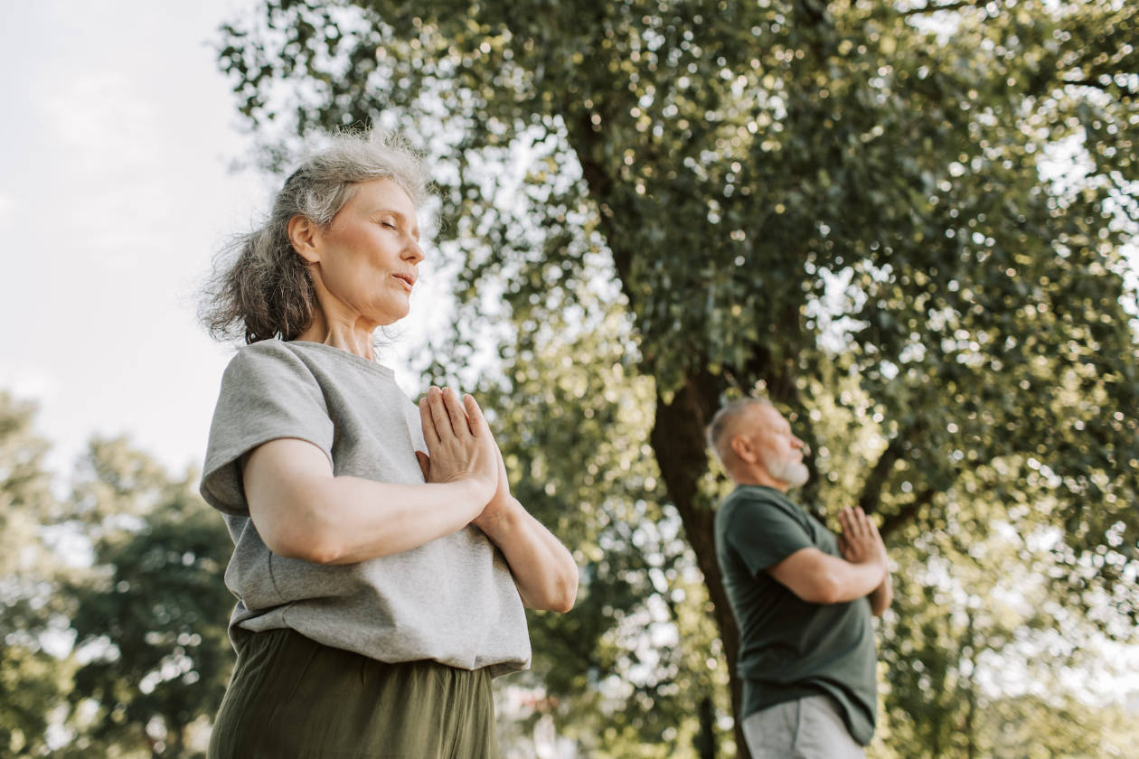 An old woman and old man doing yoga outside under the trees.