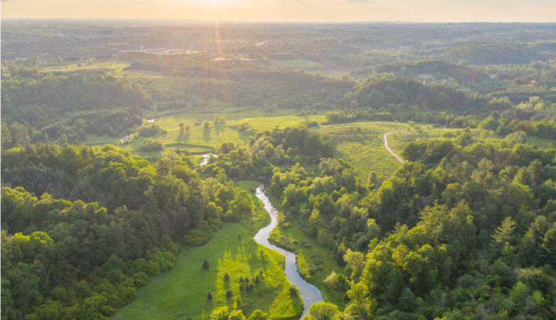 A river runs through the William Granger Greenway at Boyd Conservation Park in Ontario. The sun is setting in the distance over the lush green fields and forest.