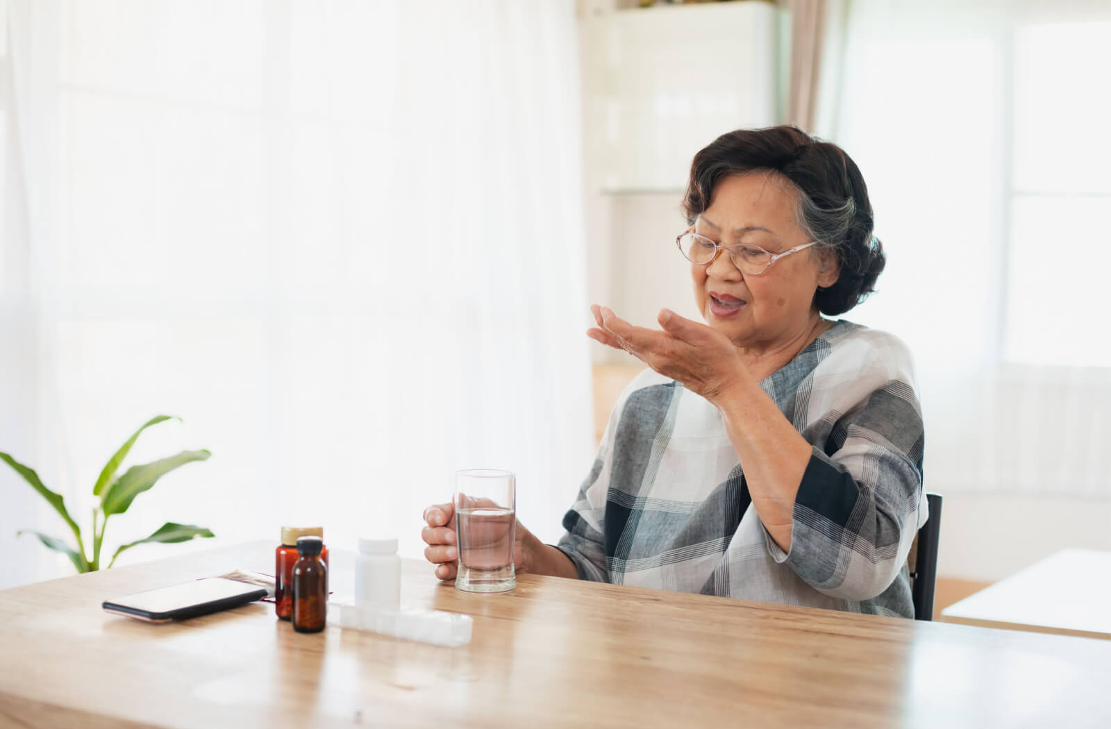 a senior woman with parkinsons sits at a kitchen table, taking medication while holding a water glass