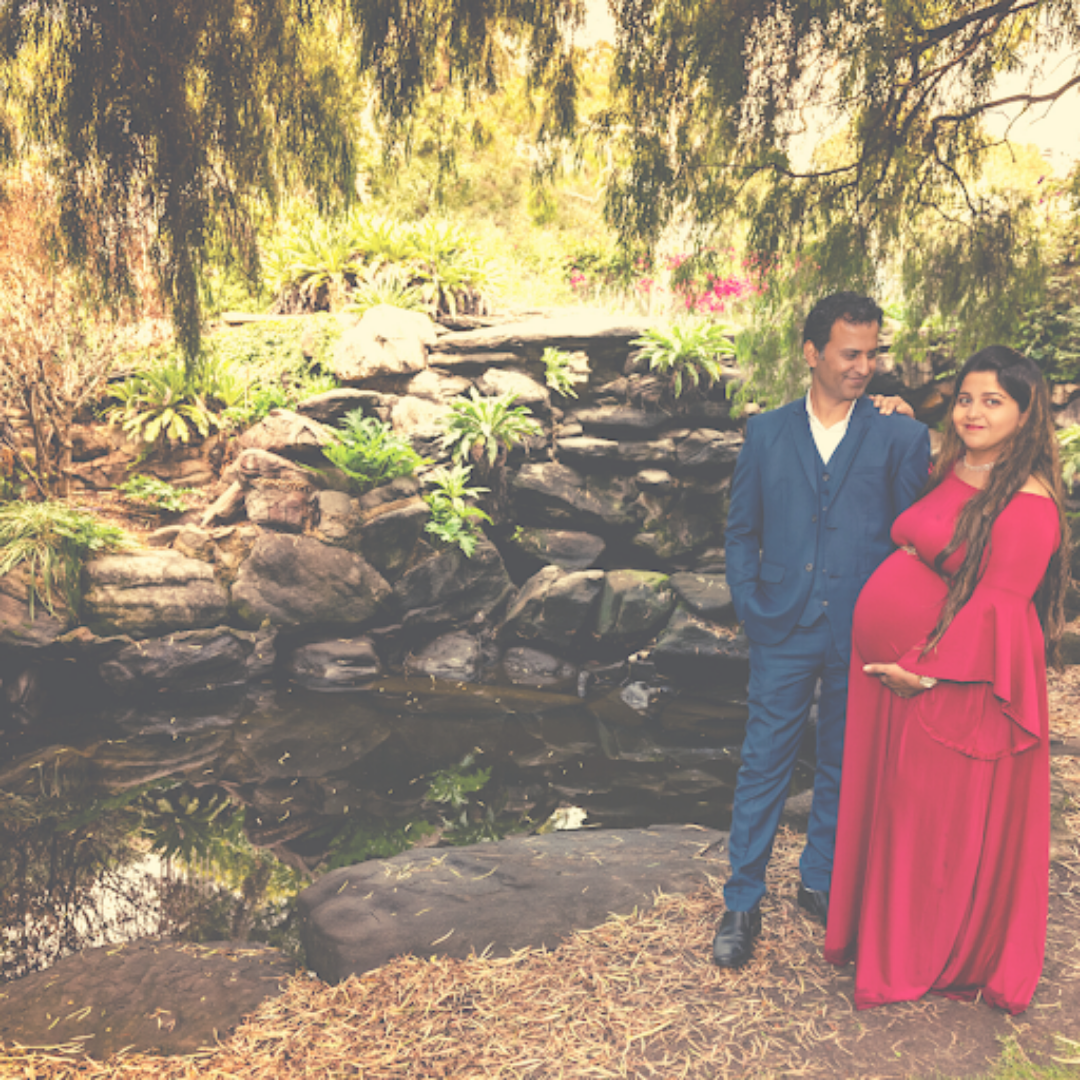 A pregnant woman in a red gown and her husband in a blue suit standing against a rocky formation