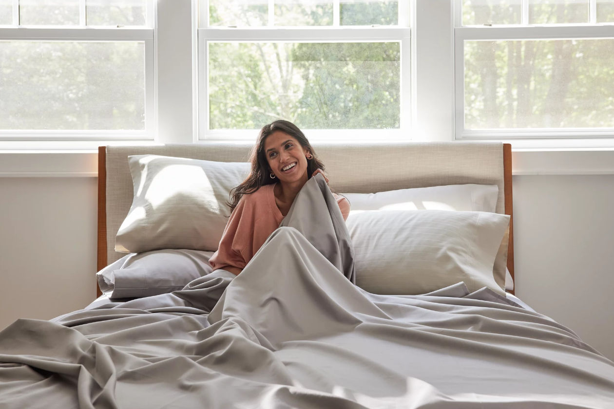 A woman smiles while sitting on a bed under gray Nestwell sheets.