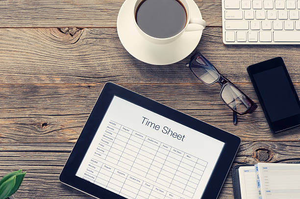 Time sheet on a digital tablet. Time sheet on a digital tablet. The tablet is on an old wooden table with a mobile phone, coffee, glasses and a keyboard. There is copy space to the right. Employment issues concept  timesheet stock pictures, royalty-free photos & images
