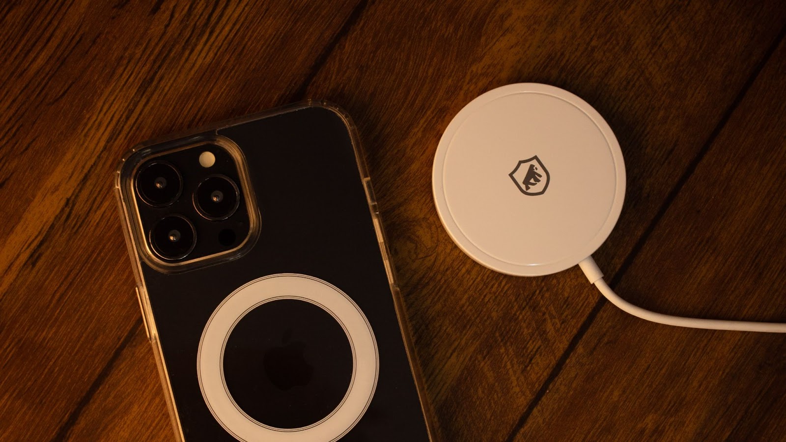 iPhone preto com capa magsafe da GShield ao lado de um carregador sem fio, em cima de uma mesa de madeira