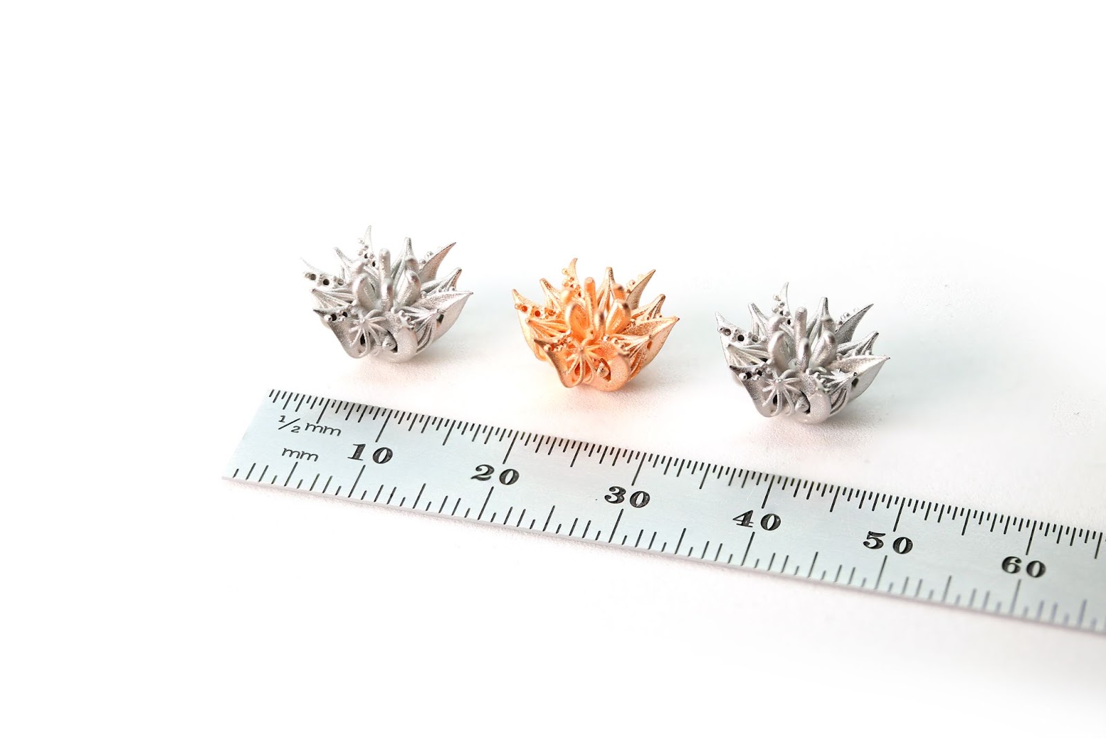 Three versions of a jewelry component, two made from stainless steels and one in copper, next to a metric ruler. Each part is roughly 10mm at their widest