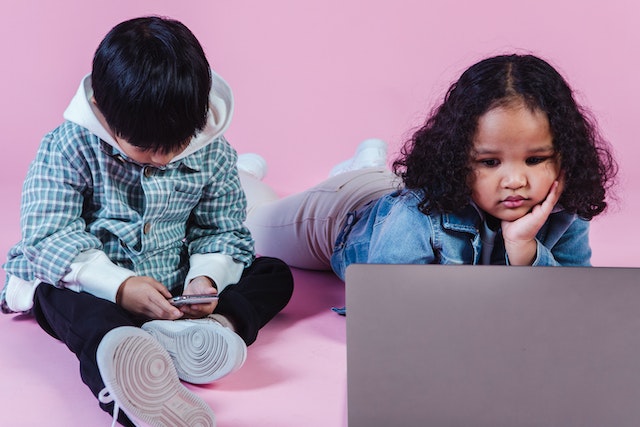 young boy sits playing on smart phone while young girl looks at laptop screen