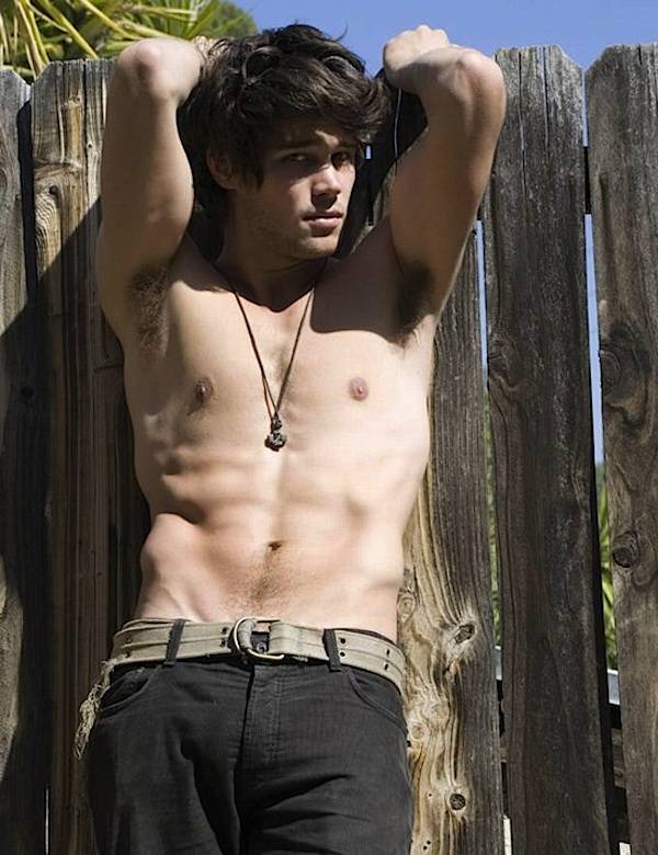 male model poses shirtless while flexing his arms against a wood fence outside