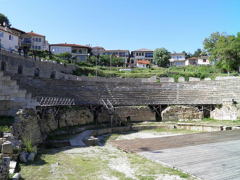 Present-day picture of an ancient Greek theater with modern houses in the background. The seating in the theater is in a semicircle around what appears to be a stage.