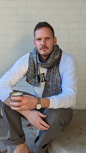 man wearing a reversible knit scarf in front of a white brick wall