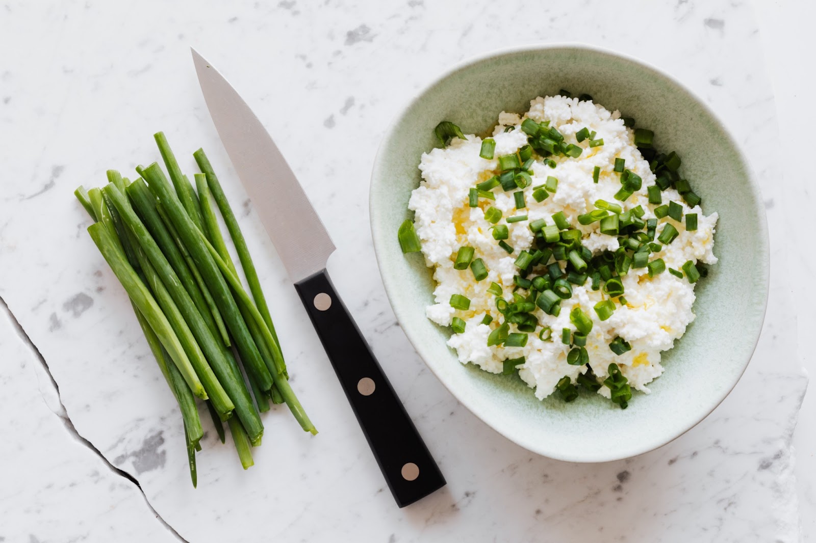 Fresh chives and a knife are pictured on a counter next to a bowl filled with cottage cheese topped with chopped fresh chives