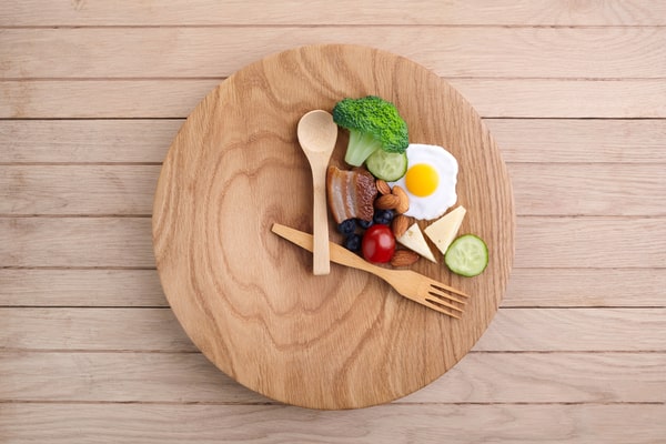 Healthy foods enclosed between a wooden spoon and fork, which are all placed on a round wooden board to resemble an analog clock