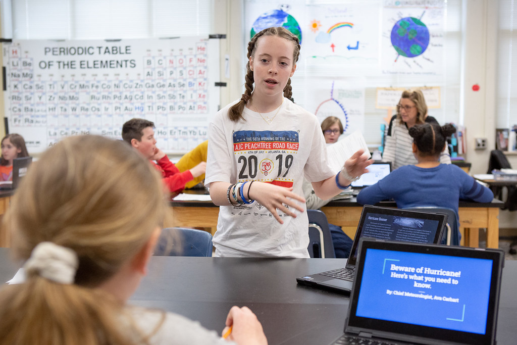 A young girl with braids stands in front of a peer at a computer. They seem to be discussing something in a science classroom. The periodic table of elements hangs on a classroom wall and other students can be seen in in the background.