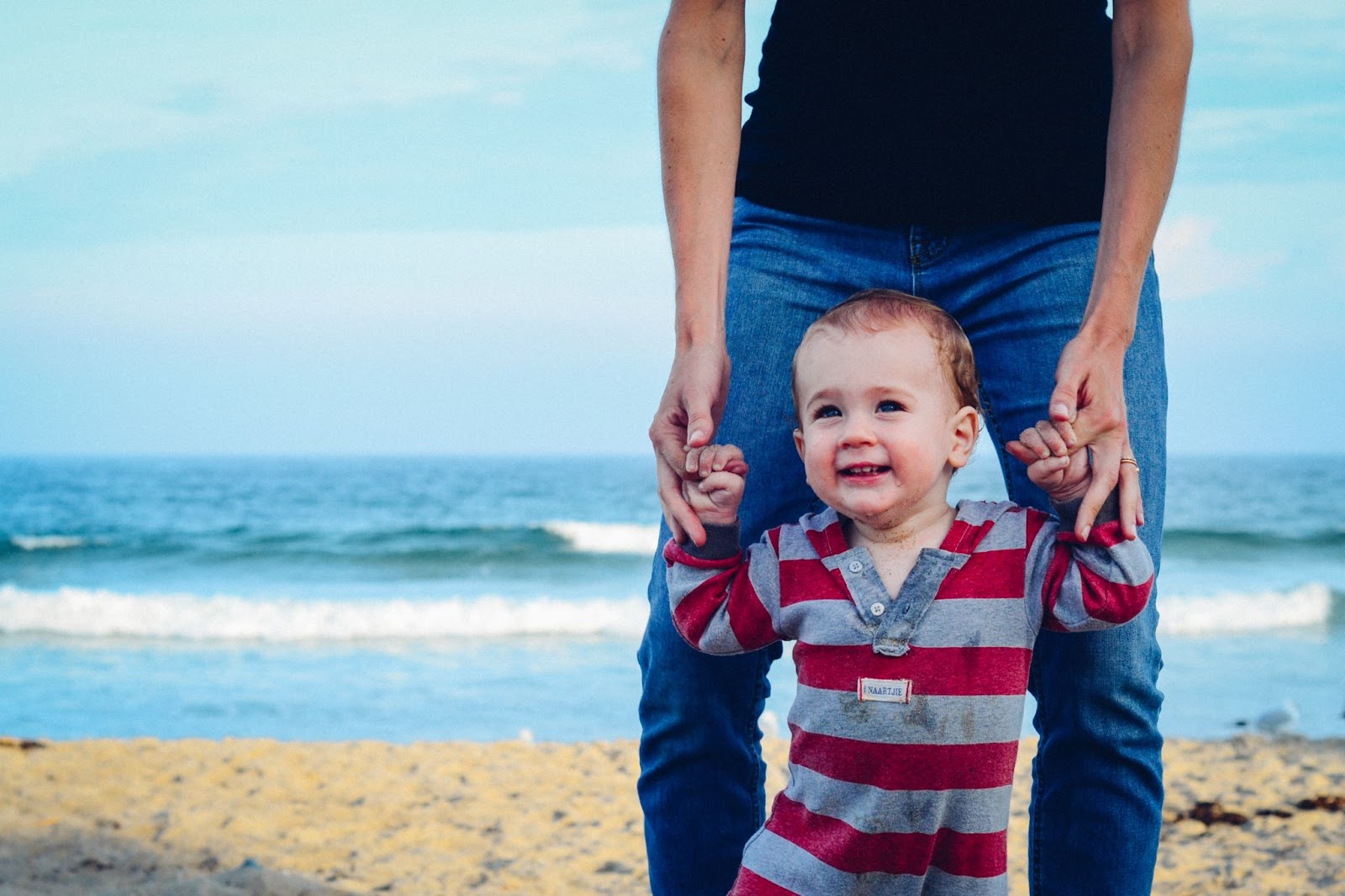 Pai segurando a mão de seu filho enquanto ele tenta andar. Ambos estão na praia