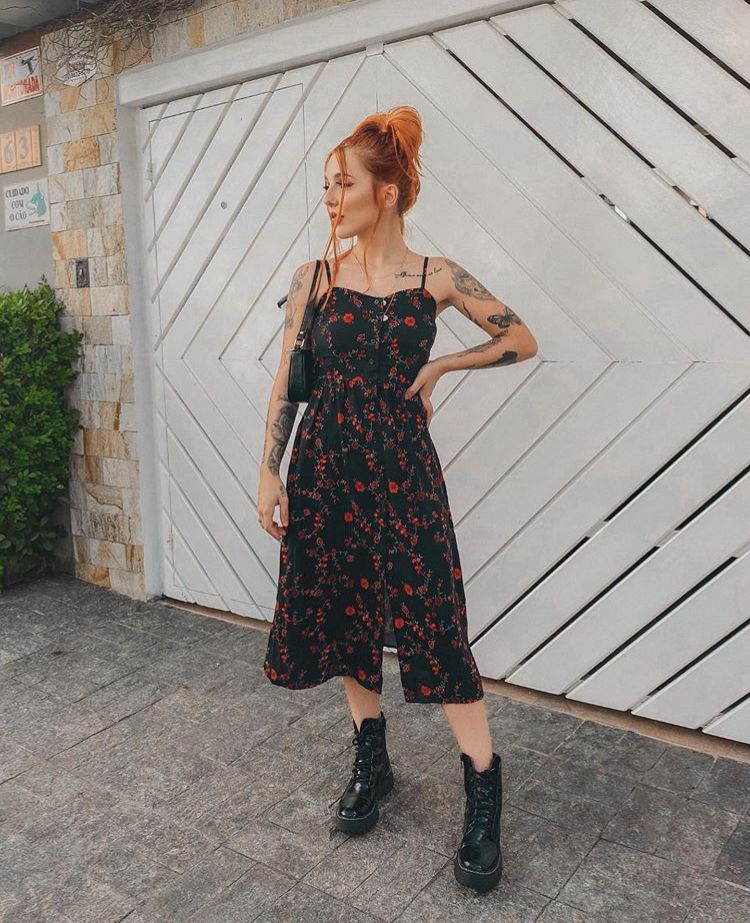 Mulher branca e ruiva, em frente a uma casa de portão de metal branco. Ela possui os braços tatuados, e apoia uma mão na cintura. Seu cabelo está preso num coque solto, e ela está vestindo um vestido médio preto com flores vermelhas, e um look com coturno clássico. 