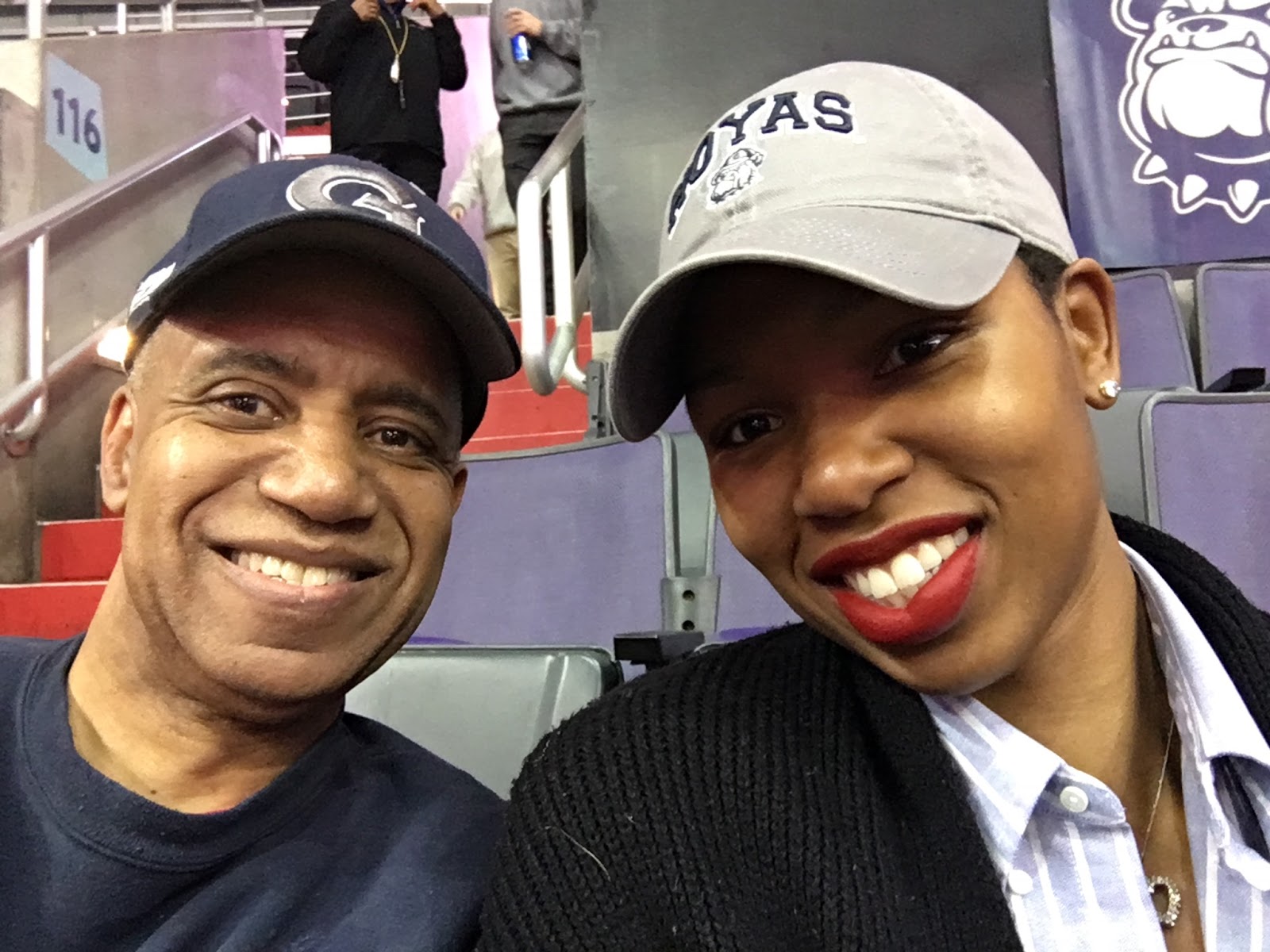 Kevin McNutt (in a blue t-shirt and blue hat with a "G") sitting next to his daughter, Monica McNutt (wearing a blue collared shirt with white stripes, a black cardigan, and a grey "Hoyas" hat) at a Georgetown University basketball game.