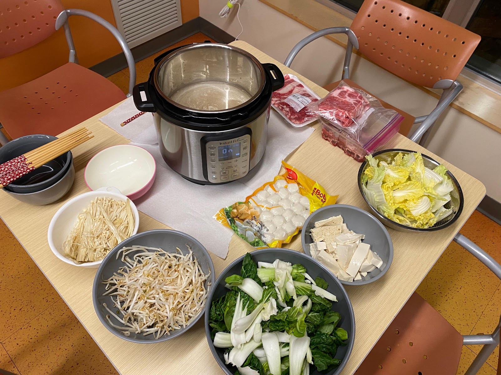 A spread of hot pot ingredients are arranged in bowls and containers on a table, surrounding an Instant Pot.