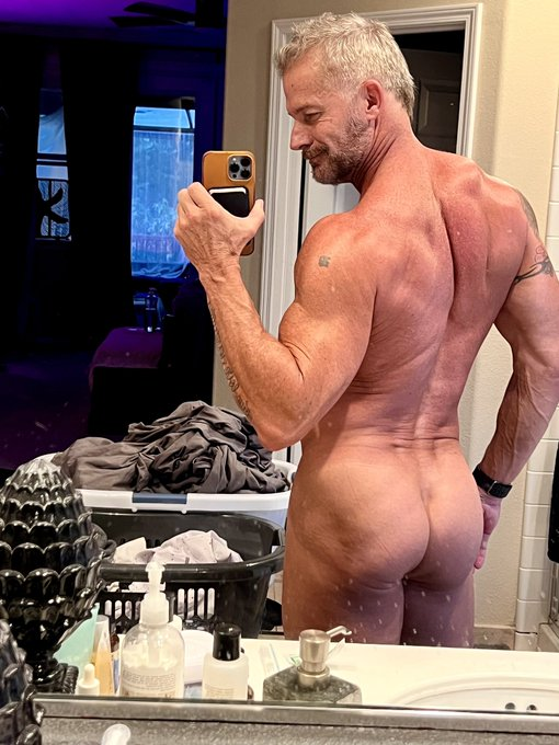 Greg Dixxon posing taking a break from folding laundry to take an iphone selfie in the mirror naked with his back facing the camera in the mirror