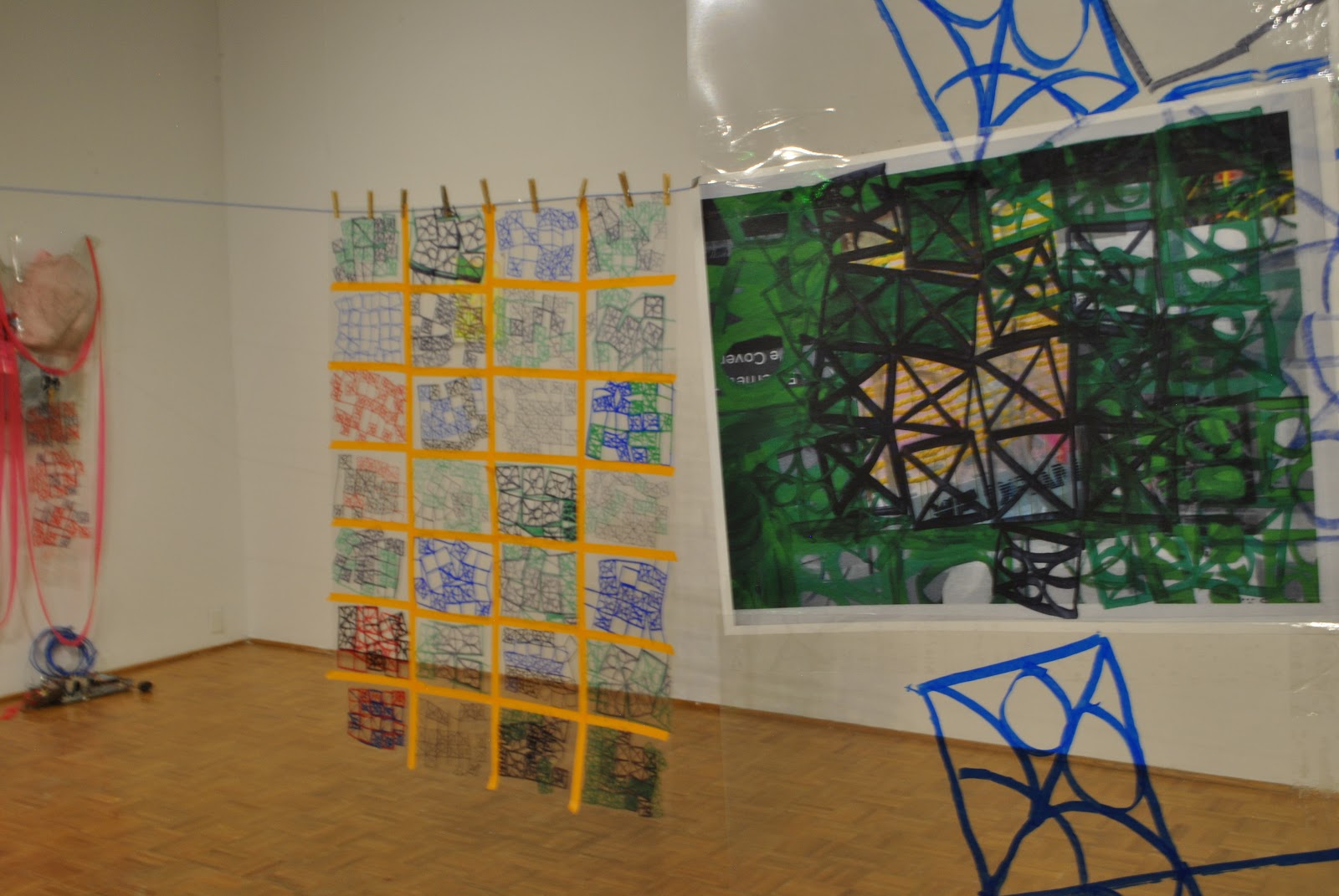 Image: Kelly Clare, enter / the net / here, 2022, Edna Carlsten Gallery. A quilt made from drawings on transparency sheets hangs from a blue rope. Drawings depict breeze block patterns with circles and squares in various formations in blue, green, red, and black marker. A grid pattern is formed by yellow tape which holds the quilt together. Image courtesy of the artist.